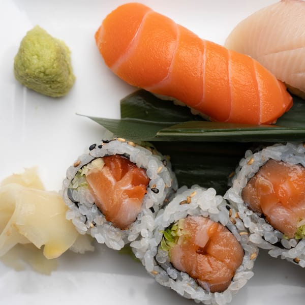 A white plate holds salmon nigiri, white fish sashimi, salmon maki rolls, wasabi, and pickled ginger.