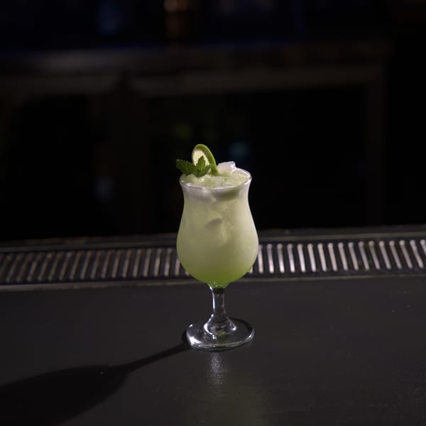 A vibrant green cocktail with ice, lime, and mint sits on a dark bar counter.