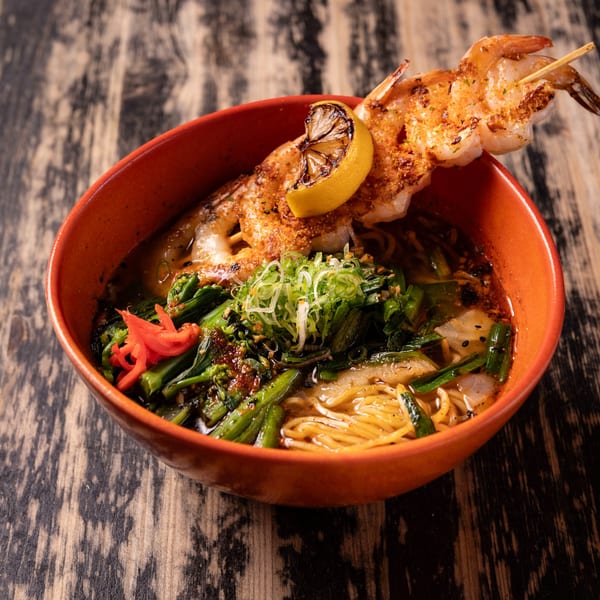 Ramen bowl with noodles, greens, and a shrimp skewer garnished with a lemon slice.
