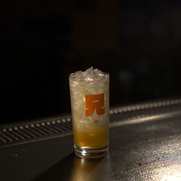 A tall glass with the "ANI" logo, filled with ice and a golden drink, sits on a dark bar.