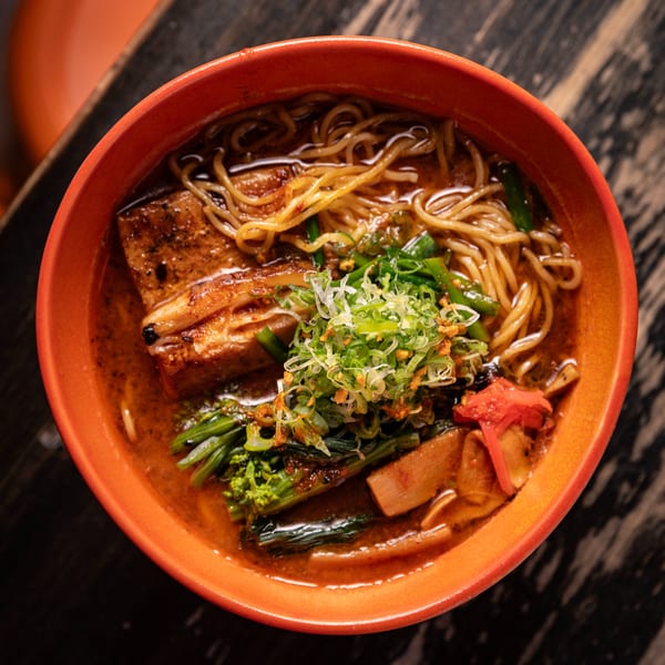 Overhead view of a vibrant orange bowl filled with ramen, pork belly, and green onions.