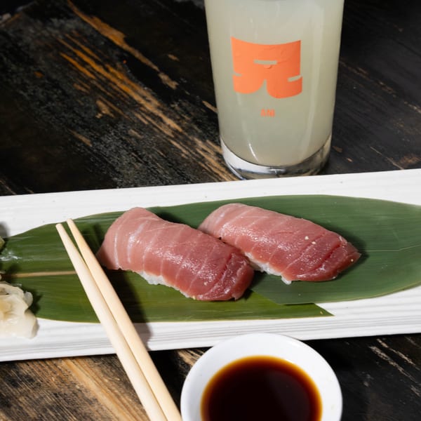 Two pieces of tuna nigiri on a banana leaf with chopsticks, soy sauce, and a lemon drink.