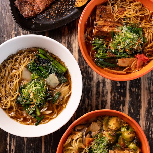 Three bowls of ramen with various toppings and a plate of grilled meat with a lemon wedge.