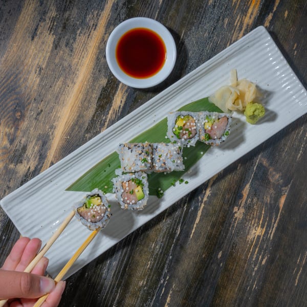 Hand using chopsticks to pick up a sushi roll from a white platter with soy sauce, ginger, and wasabi.